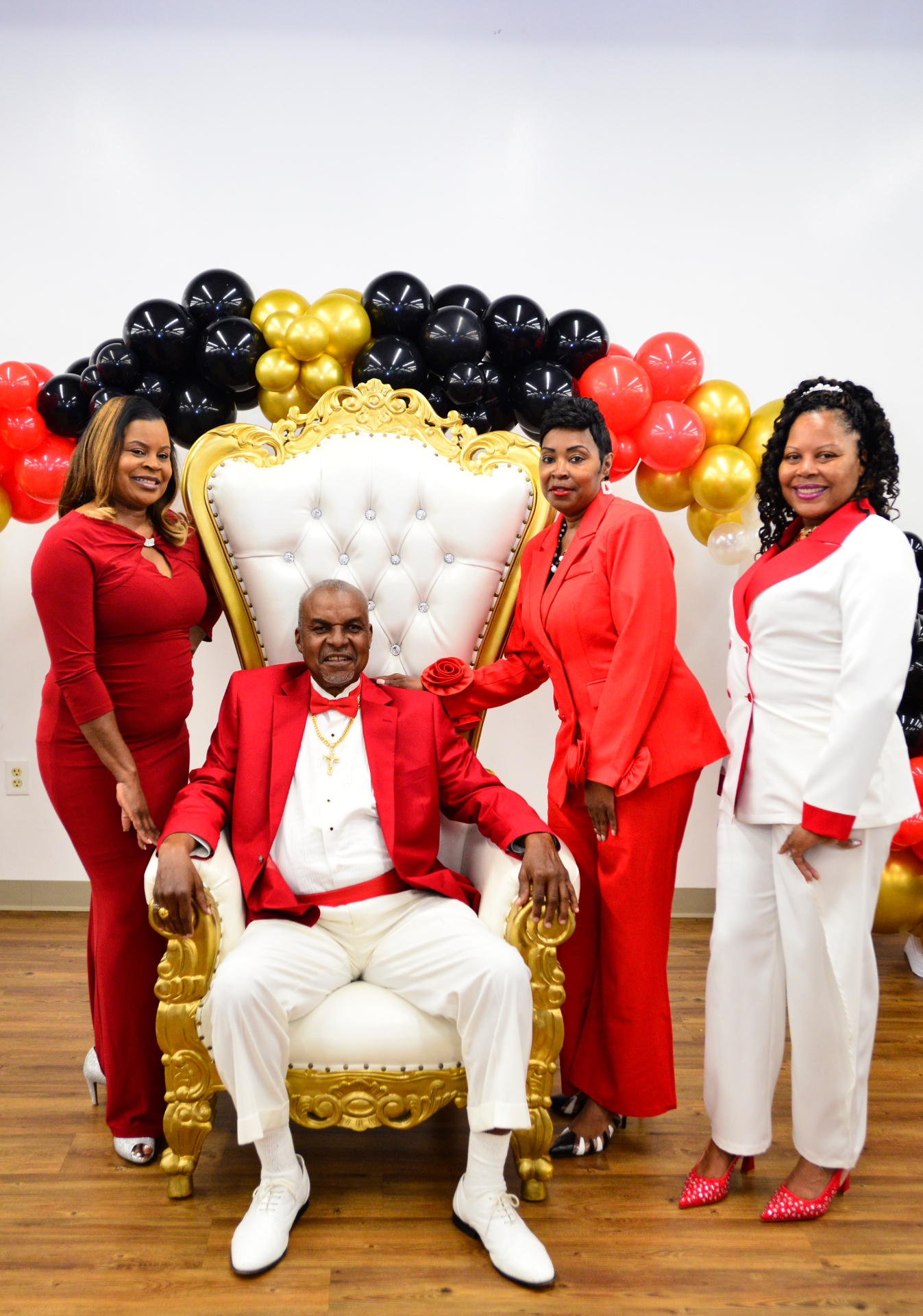 Four guests in red and white formal wear posed beside a white tufted throne chair beneath a red, black and gold balloon arch