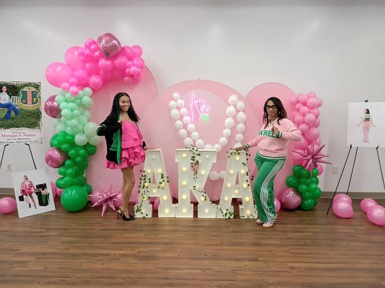Guests beside illuminated AKA letters beneath a pink and green balloon garland